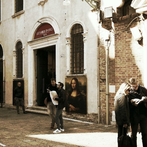 两名男子从一处博物馆入口前走过,入口处有“列奥纳多·达·芬奇”的标牌和《蒙娜丽莎》海报;另一对伴侣正在看地图。