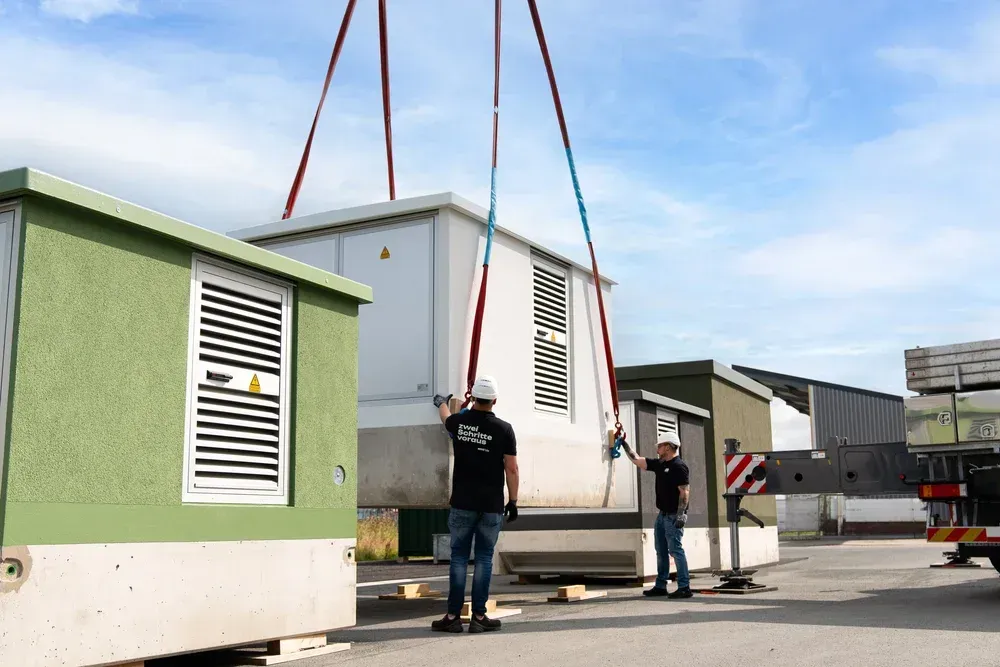 Zwei Männer beaufsichtigen, wie ein großer Transformator von einem Kran gehoben wird.