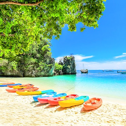 Makukulay na mga kayak na nakahanay sa buhangin na dalampasigan na may malinaw na asul na tubig, malago na berdeng mga puno, at mabatong bangin sa likuran.