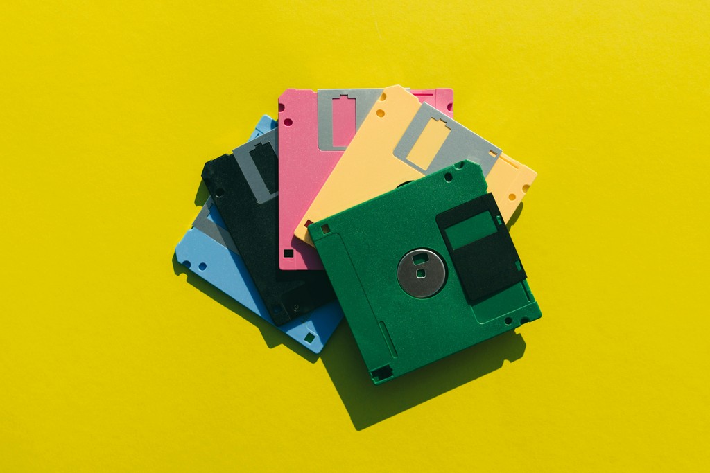 A colorful stack of vintage floppy disks in various colors (pink, yellow, green, blue, black, and gray) sits on a bright yellow background, highlighting retro computer storage technology.