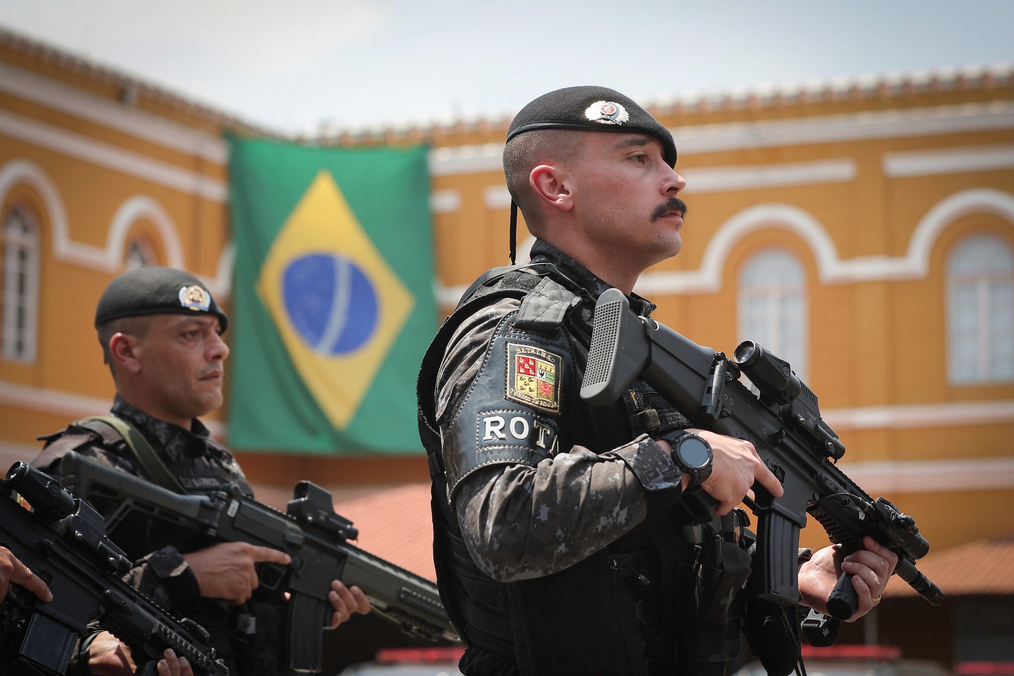 policial da ROTA segurando uma arma na mao com uma bandeira do brasil ao fundo