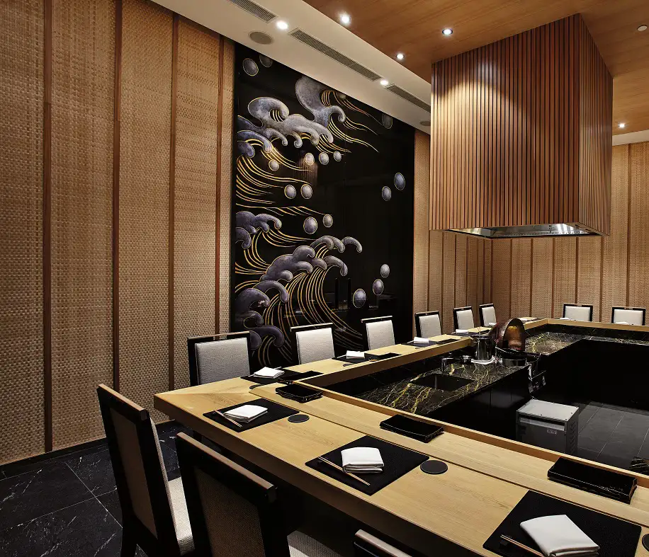 A modern, upscale Japanese restaurant interior featuring a long, elegant wooden dining table with place settings, contemporary black and grey chairs, a striking wall art of waves and spheres, and ambient overhead lighting enhancing the sophisticated atmosphere.