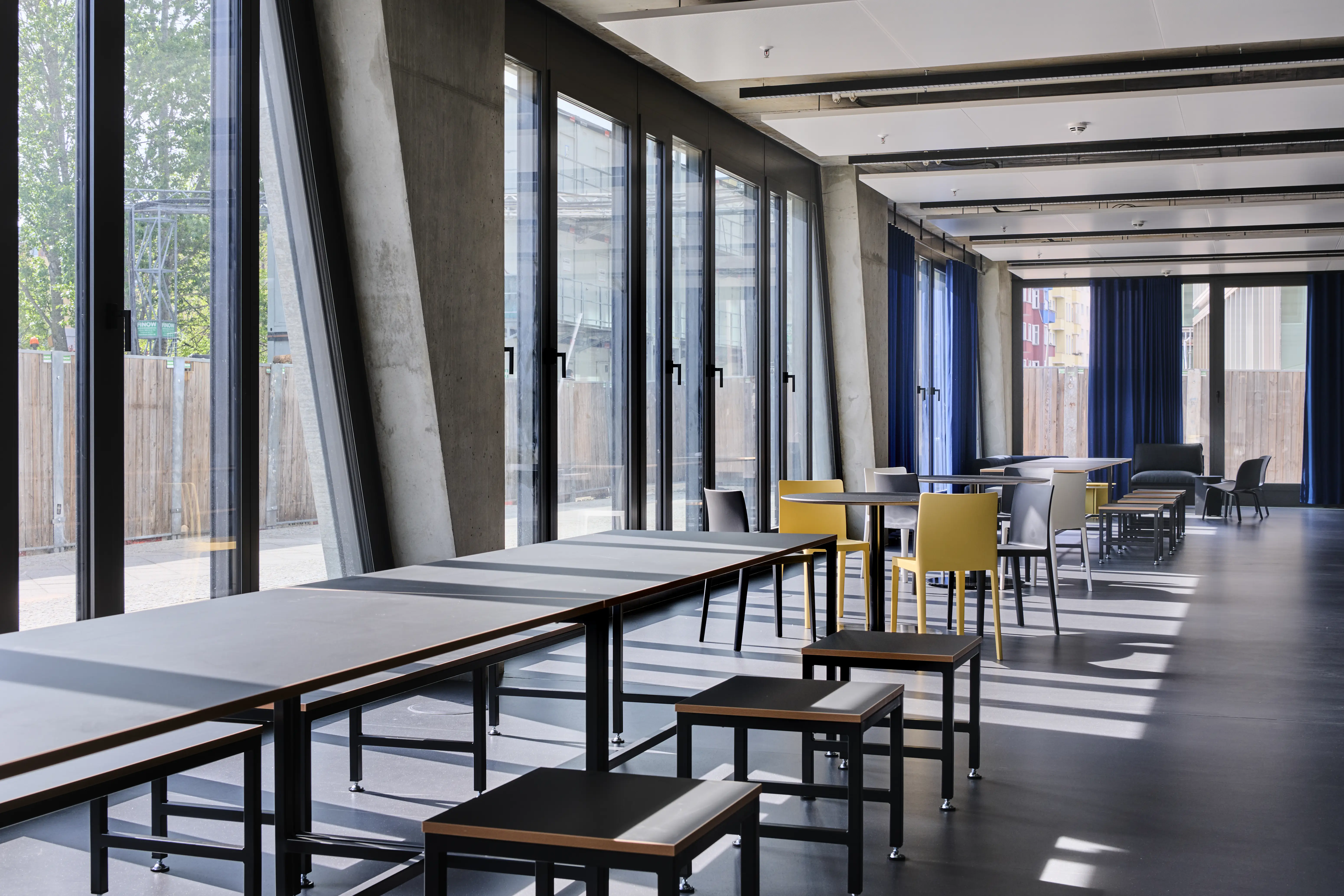 Canteen at ENPAL along slanted concrete columns and floor-to-ceiling windows, with black tables, yellow and black chairs, low black stools and royal-blue curtains.