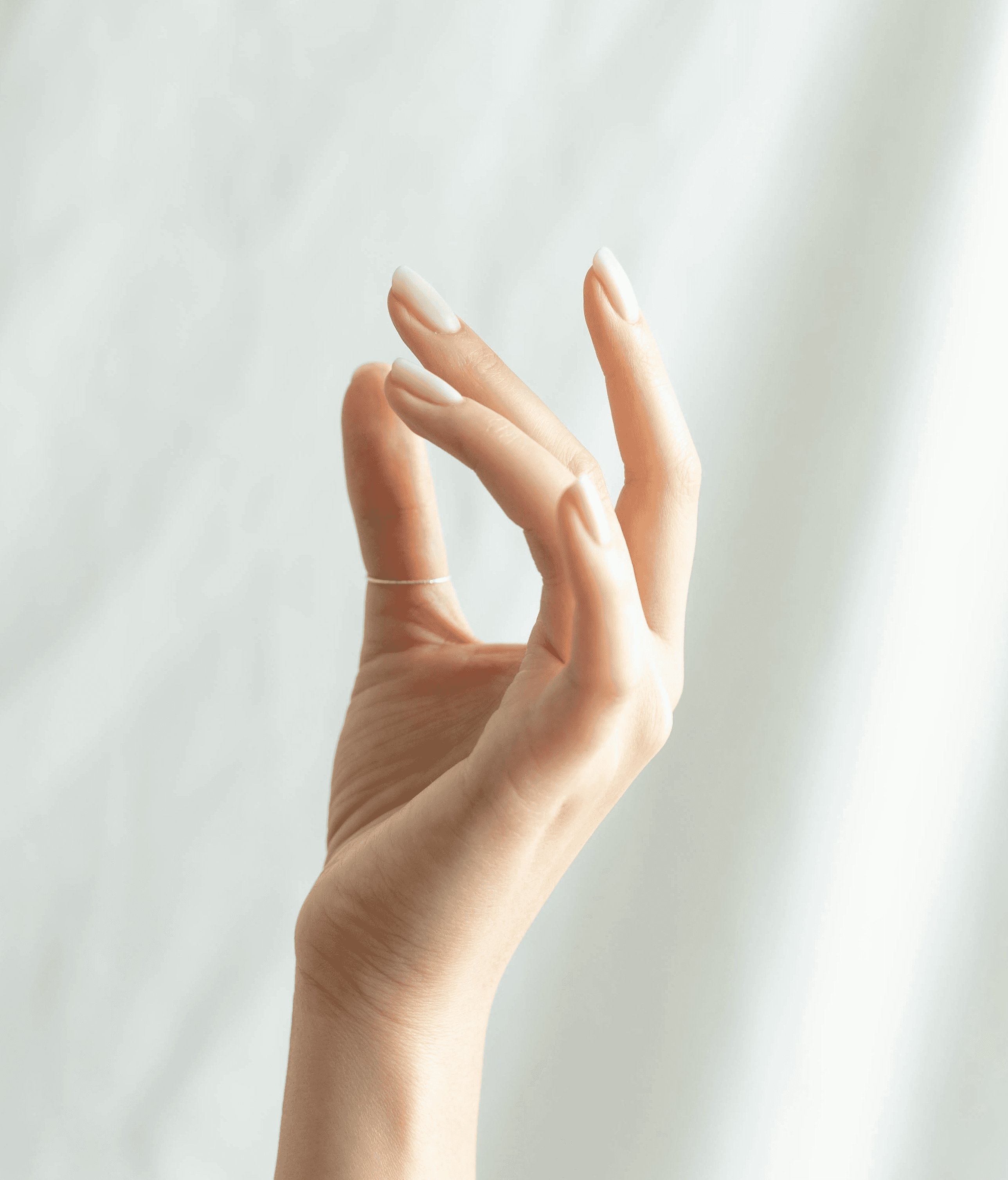 A soft, minimalist shot of a hand with a thin silver ring posed elegantly against a draped white fabric background.