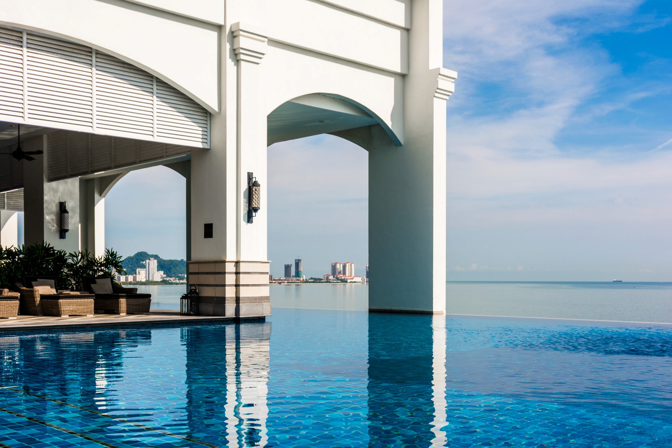 An infinity pool at a white coastal resort overlooks a blue ocean under a bright sky with light clouds.