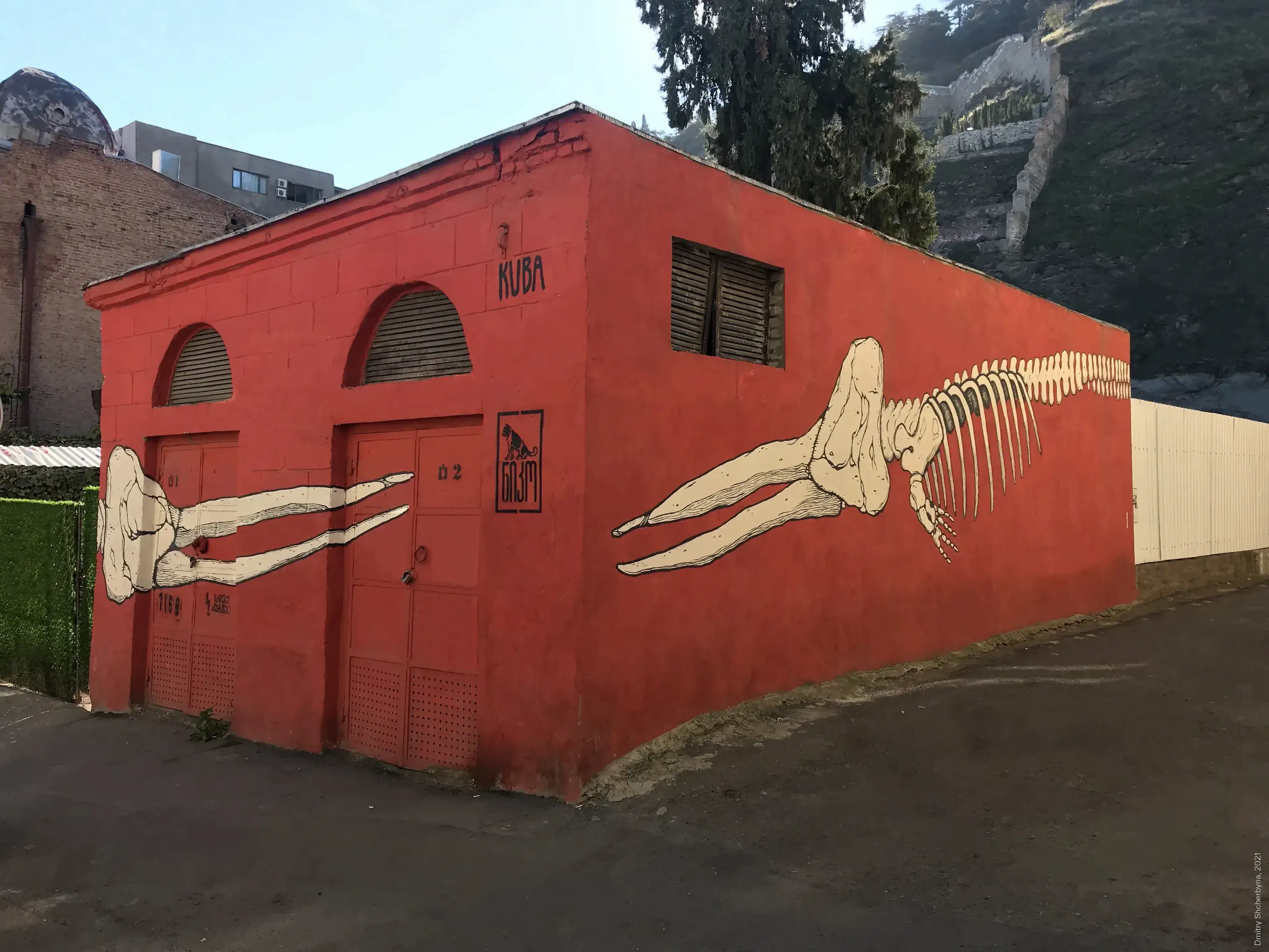 A mural wraps around the corner of the red building, depicting a large animal skeleton in cream and black lines that stretch across both walls. Its legs extend past the doorway.