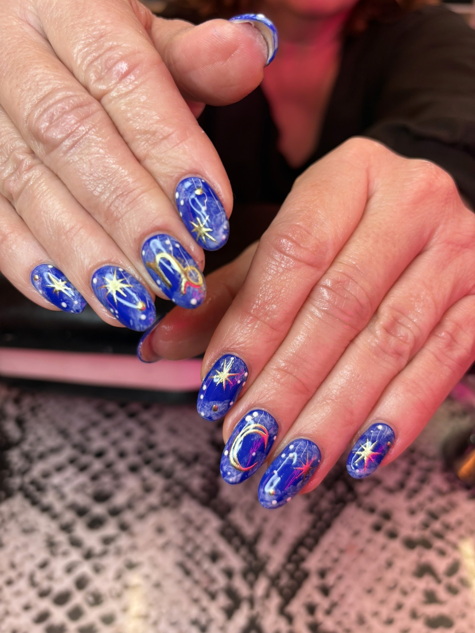 Close-up of almond-shaped nails featuring a deep blue galaxy base, hand-painted gold stars, and a golden Capricorn zodiac symbol.