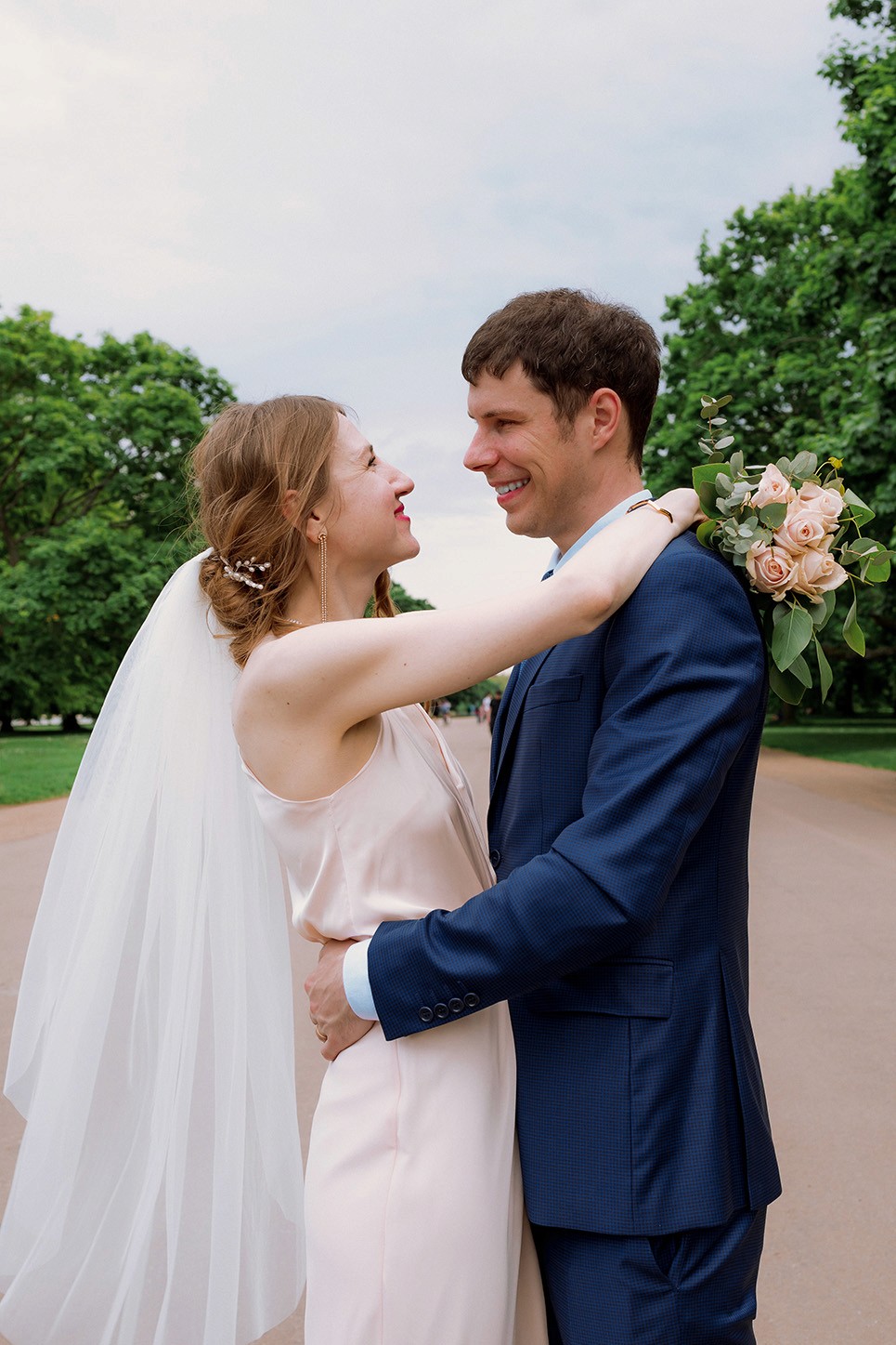 Romantic outdoor wedding portrait of bride and groom embracing on a tree-lined pathway with bouquet and veil details in soft natural light
