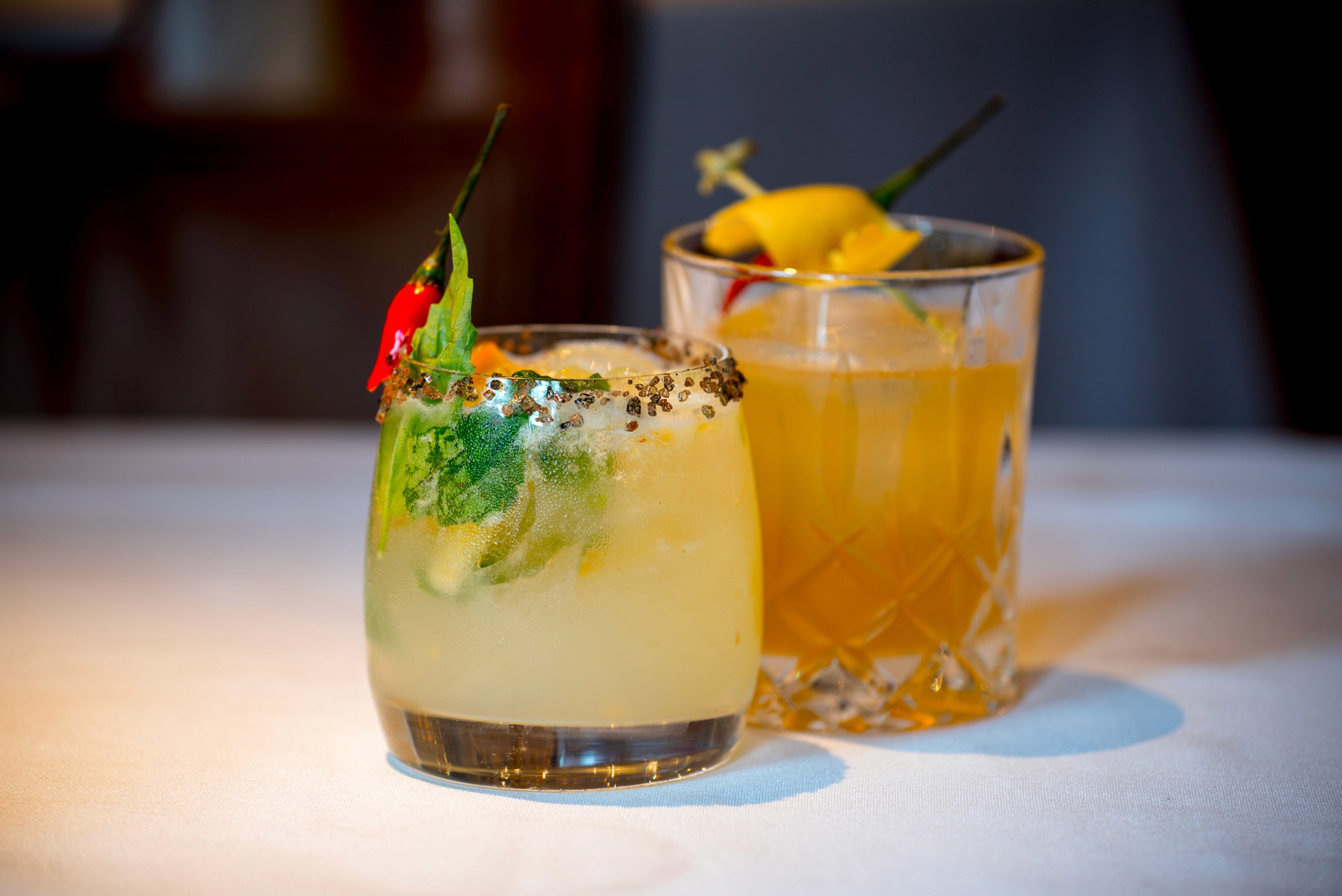 Two cocktails in glasses on a white table; one with a chili pepper and lime, the other with an orange slice.