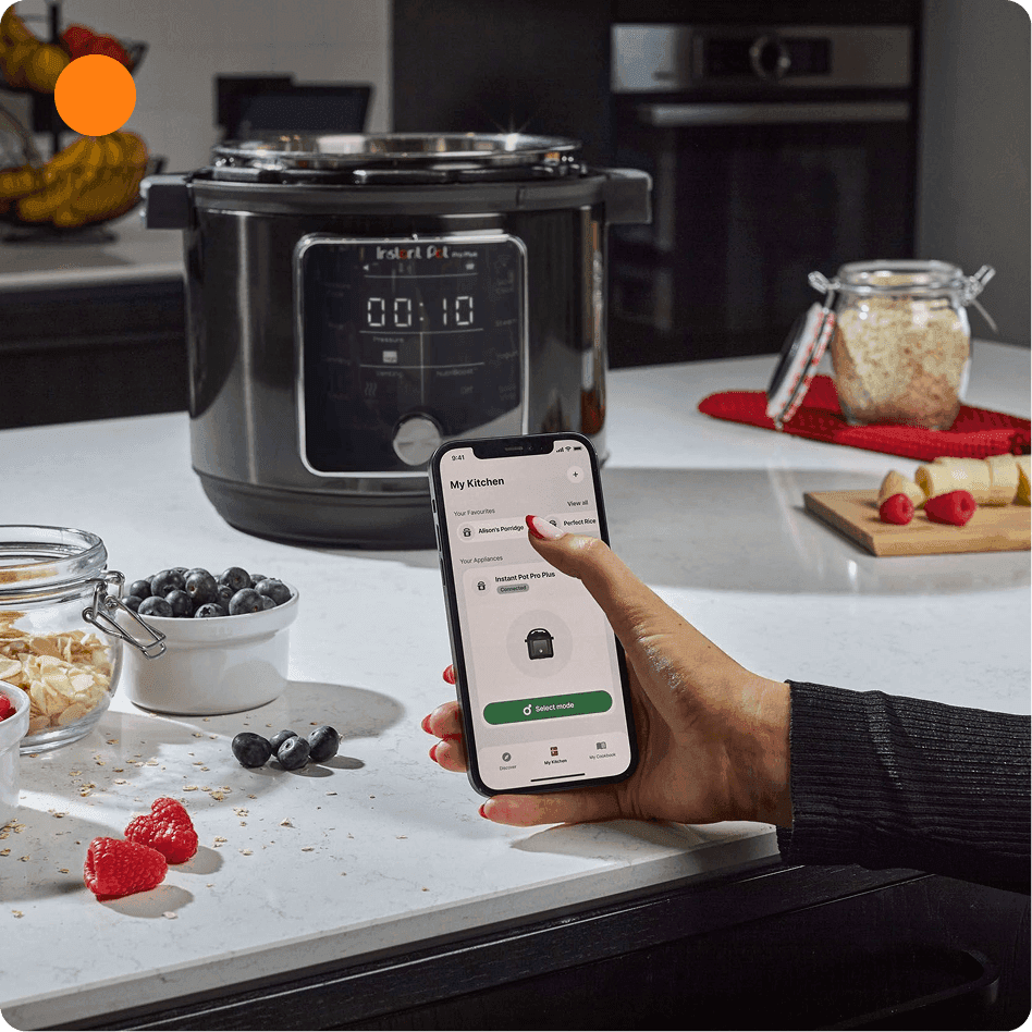 Hand holds a smartphone controlling a multicooker with a timer display while ingredients like berries and oats sit on the counter