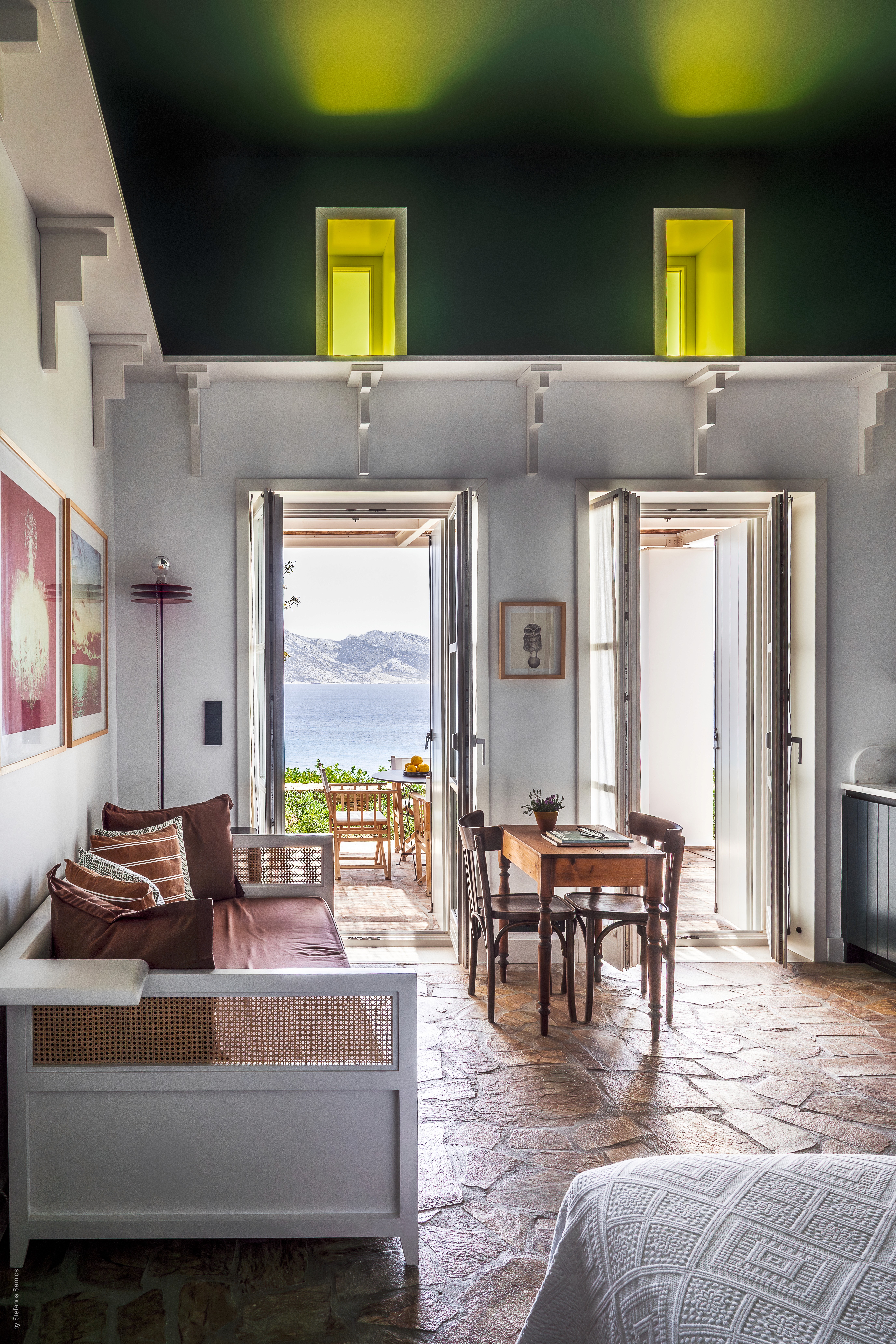 A living room of a Greek house with a white sofa, green ceiling and yellow windows looking out onto the ocean