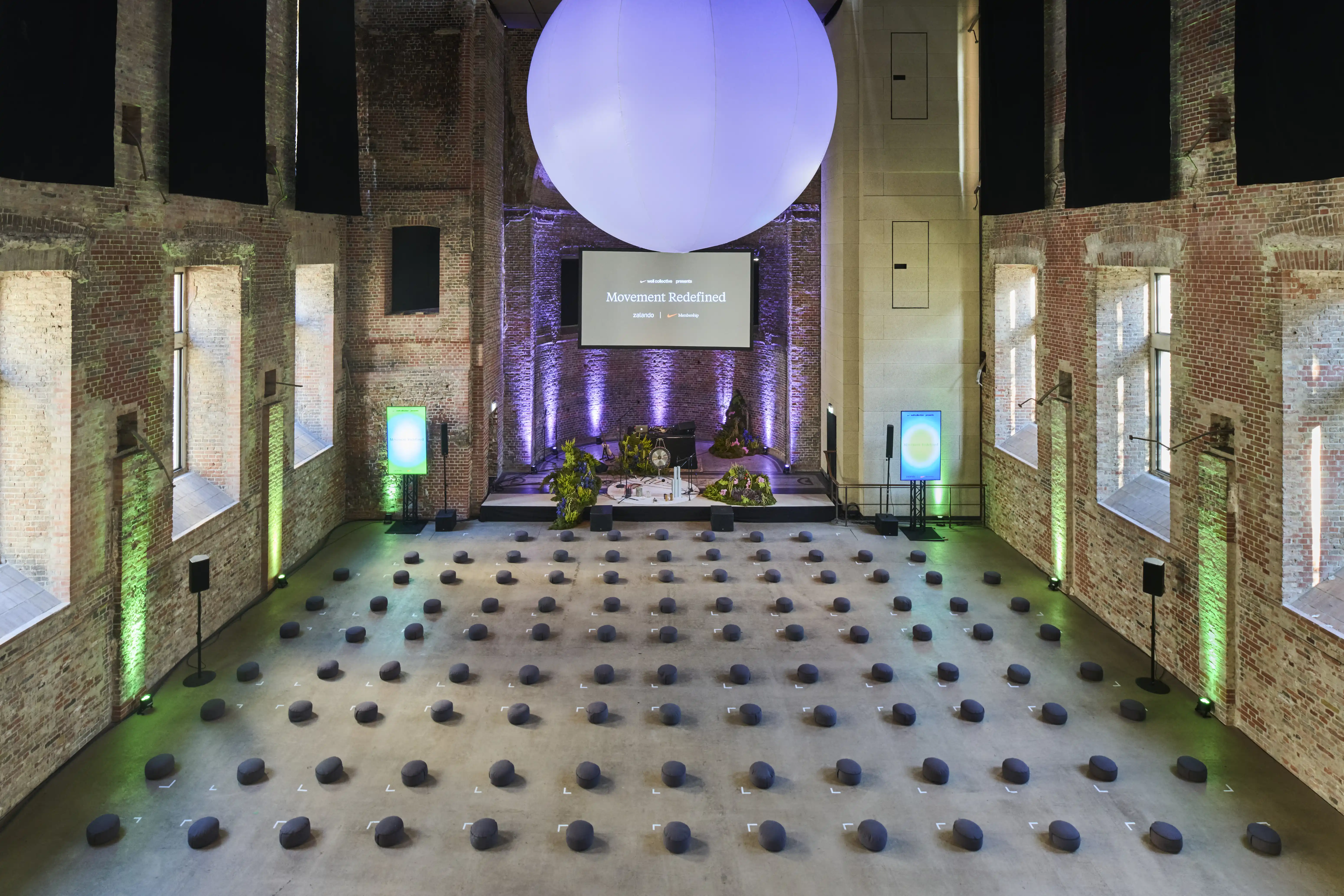 Brick event hall with rows of meditation cushions, stage and a large balloon sphere overhead.