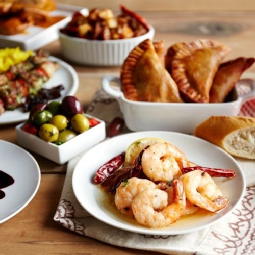 Eine Vielzahl von Gerichten, darunter Empanadas, Garnelen mit Paprika, Spieße, Oliven und Brot auf einem Holztisch.