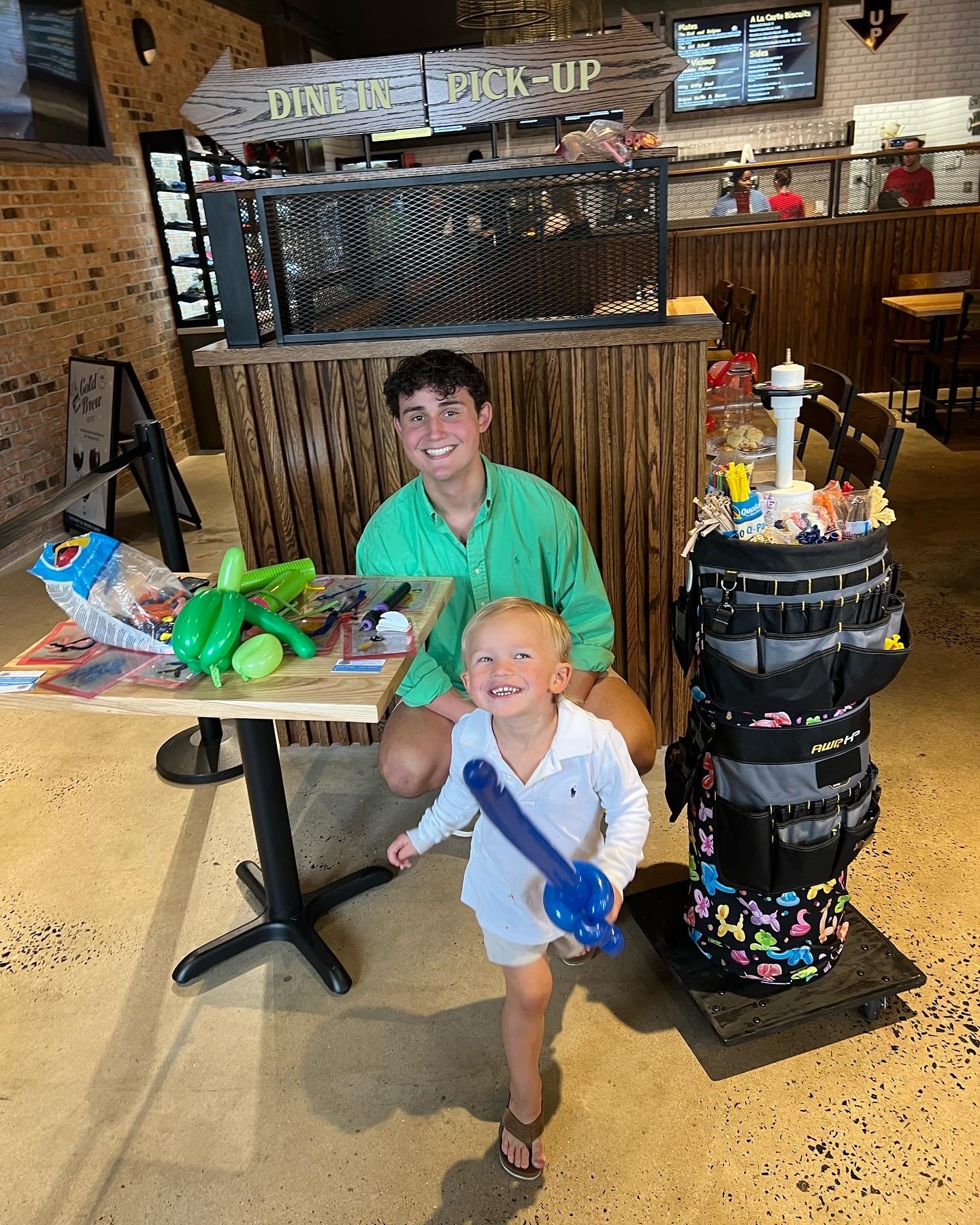 Balloon artist seated at a table inside Vicious Biscuit, smiling alongside a cheerful young boy holding a blue balloon sword. The setup includes colorful balloon creations and supplies, with the warm interior of the restaurant in the background.