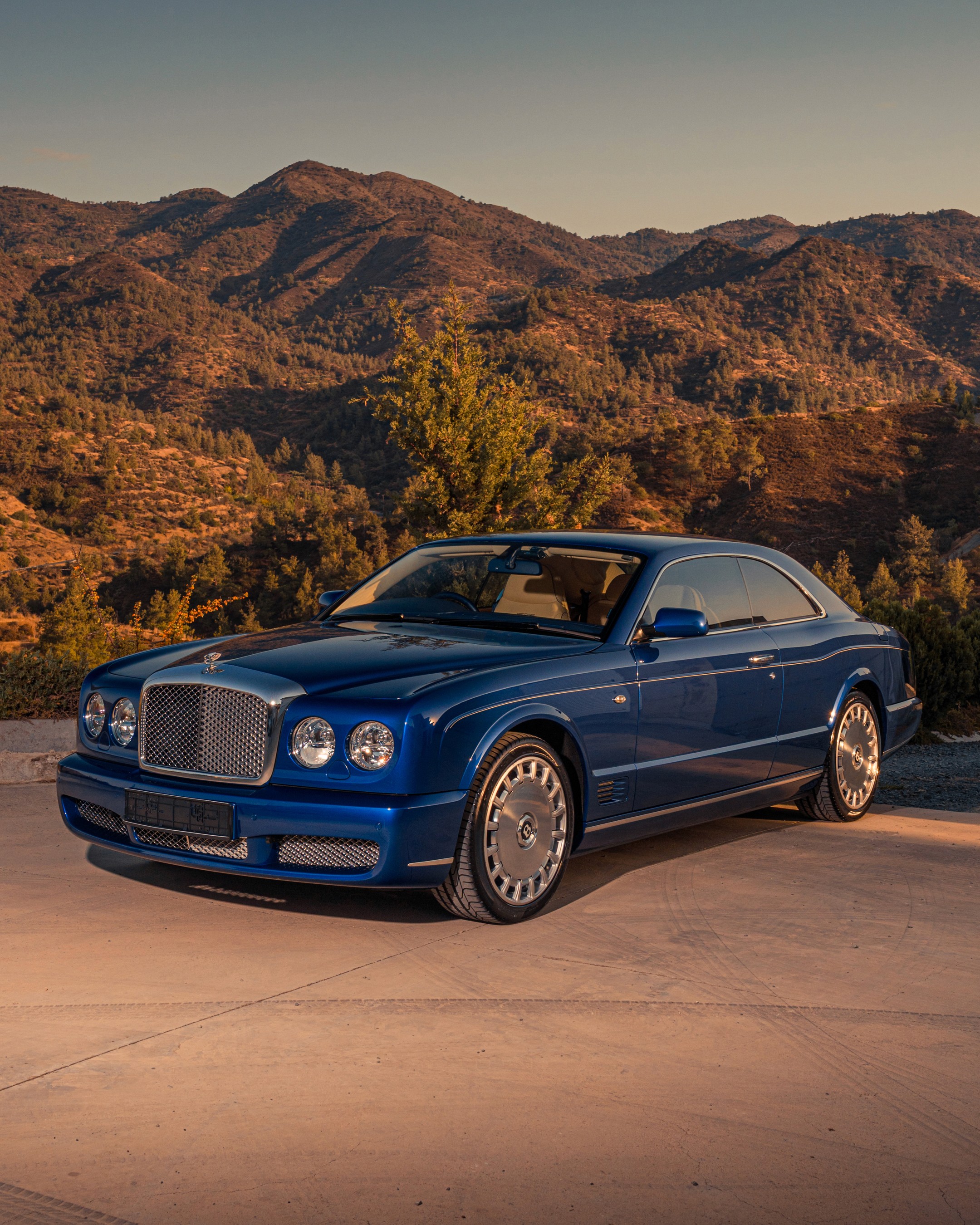 Bentley Brooklands coupé in Moroccan Blue photographed from the front three-quarter, chrome mesh grille and quad round headlamps facing forward, against the tree-covered foothills of Troodos, Cyprus in warm evening light.