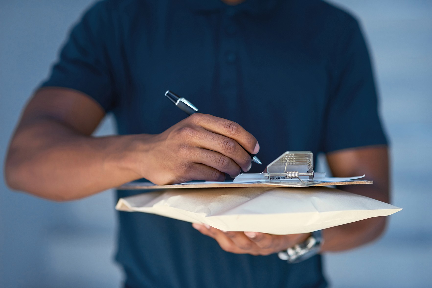 close up view of large brown skinned hands hovering a pen over a clipboard, signing for papers 