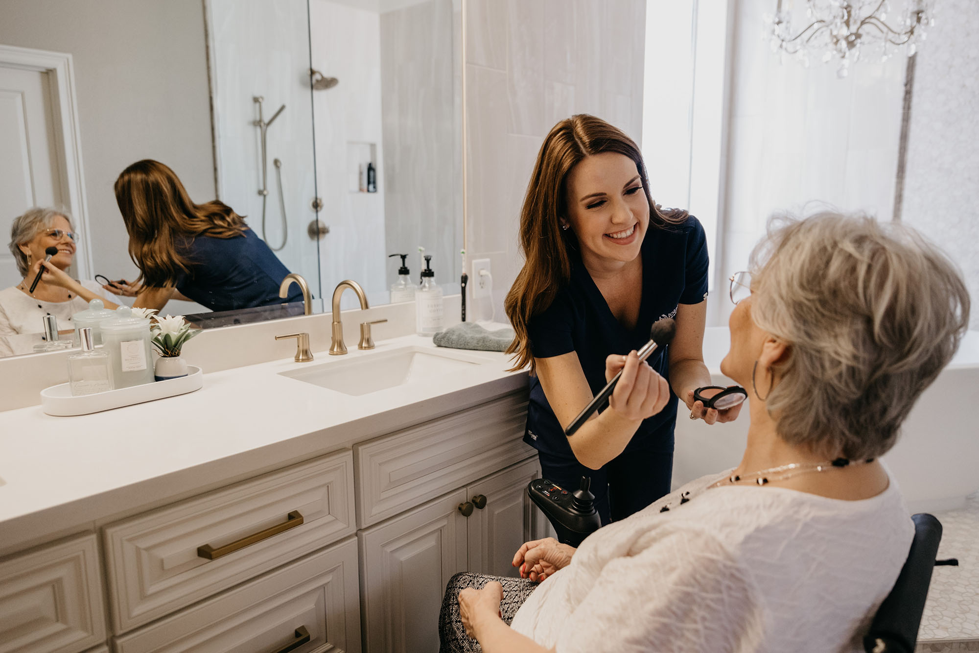 A younger woman applying makeup to an older woman sitting in a wheelchair in front of a bathroom vanity.