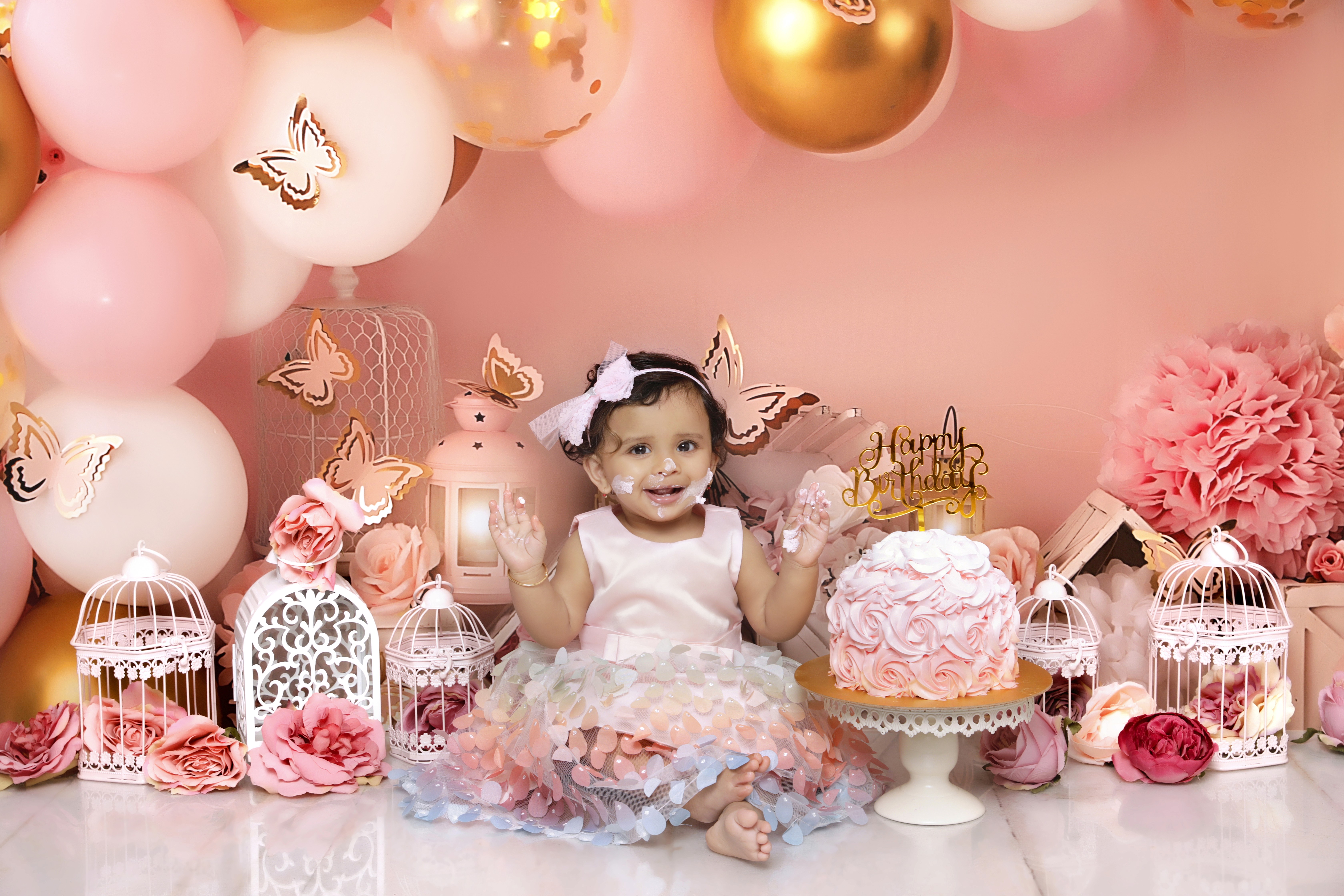 Smiling baby girl with cake frosting on her face surrounded by pink floral and butterfly decor during a first birthday cake smash.