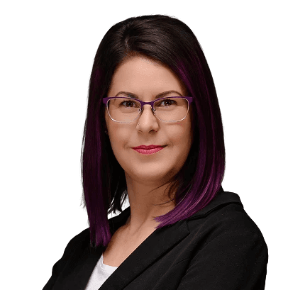 Lilás hajú nő portréja lila szemüvegben, sötét blézerben, kék háttérrel.