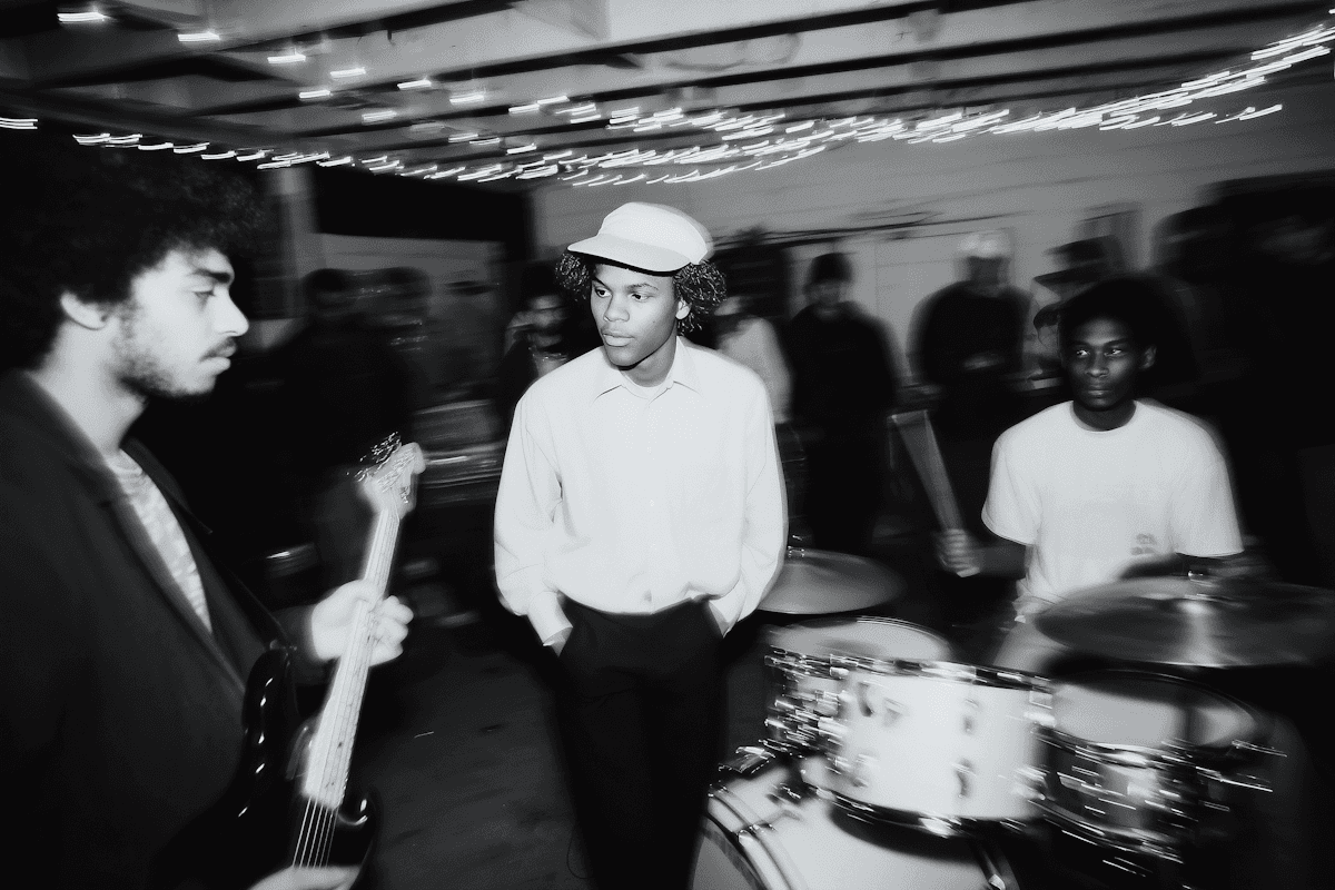 Black-and-white photo of a band rehearsal with guitarist, drummer, and people gathered under string lights.