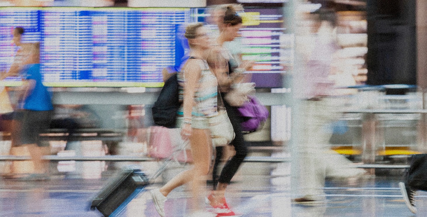 A motion blur image of people walking in front of the time board at an airport with luggage.