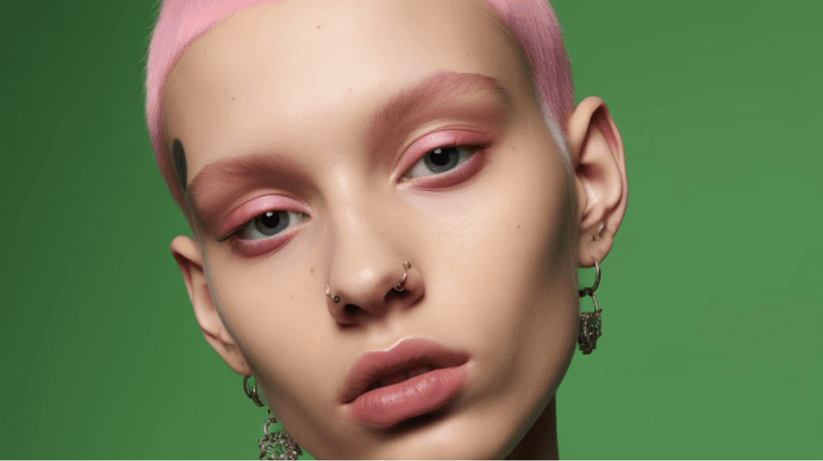 Close-up portrait of a model with pastel pink buzz-cut hair, soft pink eyeshadow, and multiple facial piercings, set against a vibrant green background.