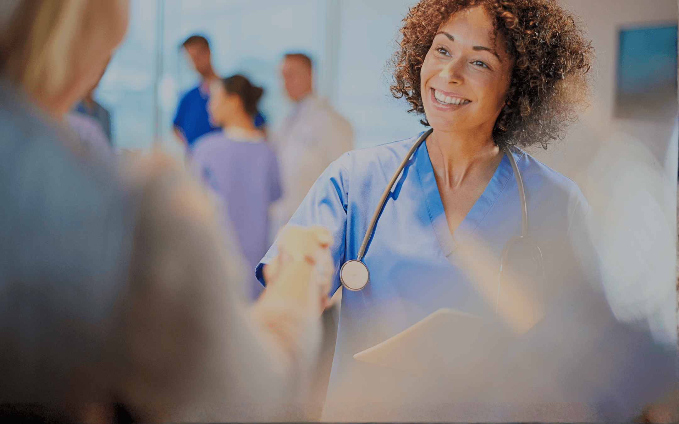 Smiling nurse shakes a patient's hand. Other medical professionals in scrubs stand in the background.