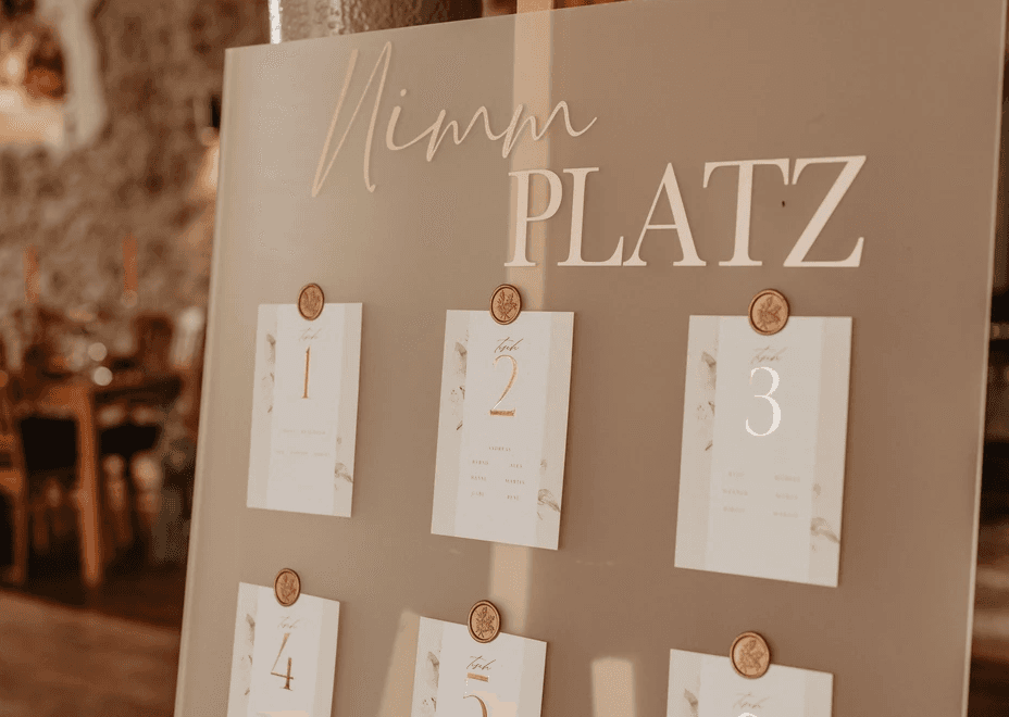 Eleganter und moderner Sitzplan in matter Acryl-Optik mit dem Titel 'Nimm Platz' und nummerierten Kärtchen mit goldenen Wachssiegeln.