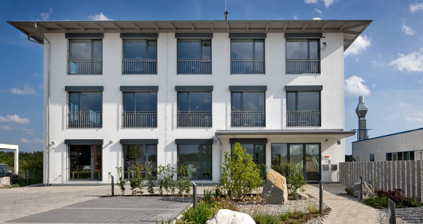 Modernes weißes Bürogebäude mit großflächigen Fenstern, Balkonen und einer gepflegten Außenanlage.