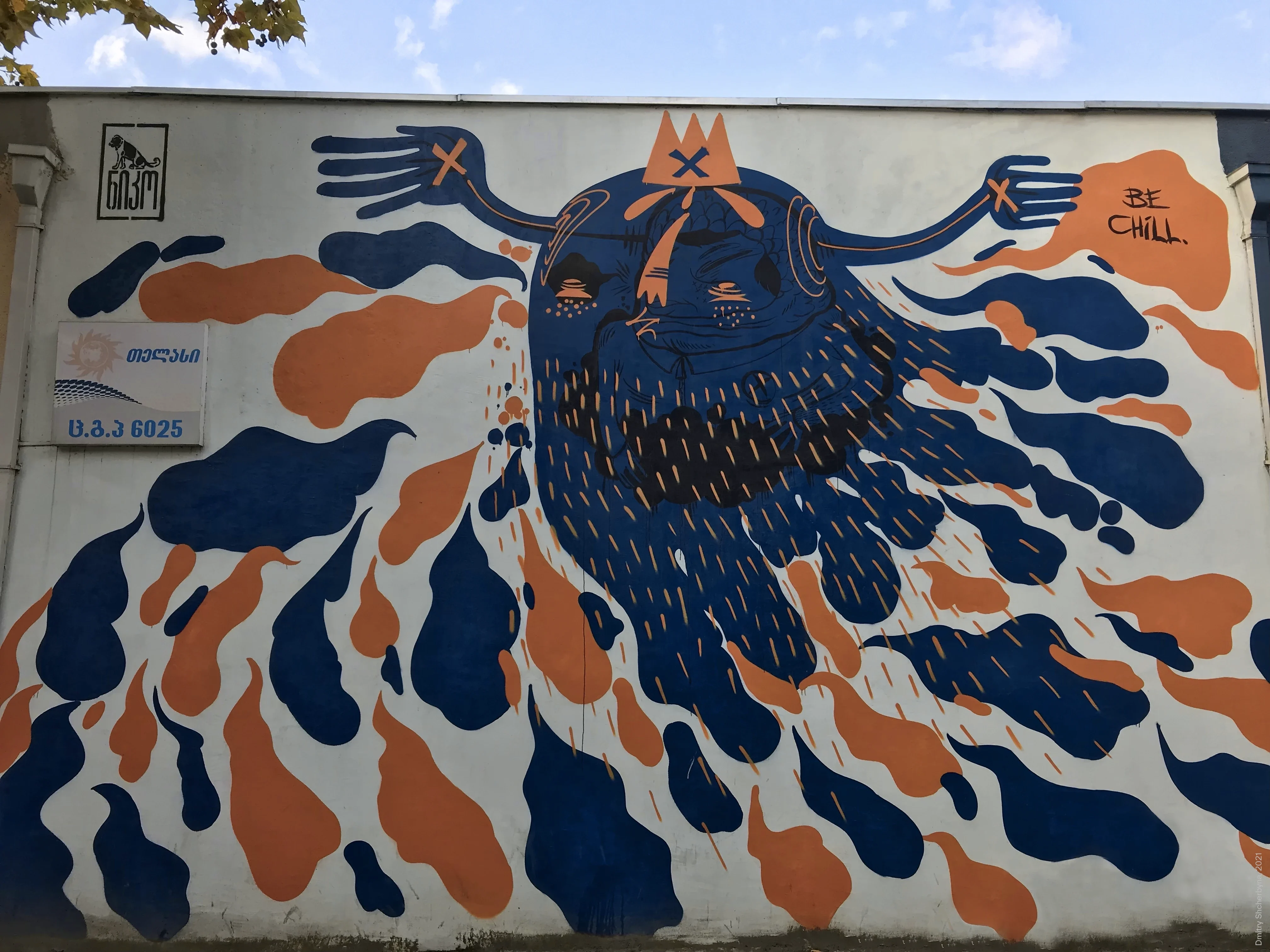 A graphic mural featuring a round-faced bearded figure wearing a small orange crown, with navy blue and orange teardrop-shaped splashes radiating outward across a white wall. The word "BE CHILL" is written in large letters.