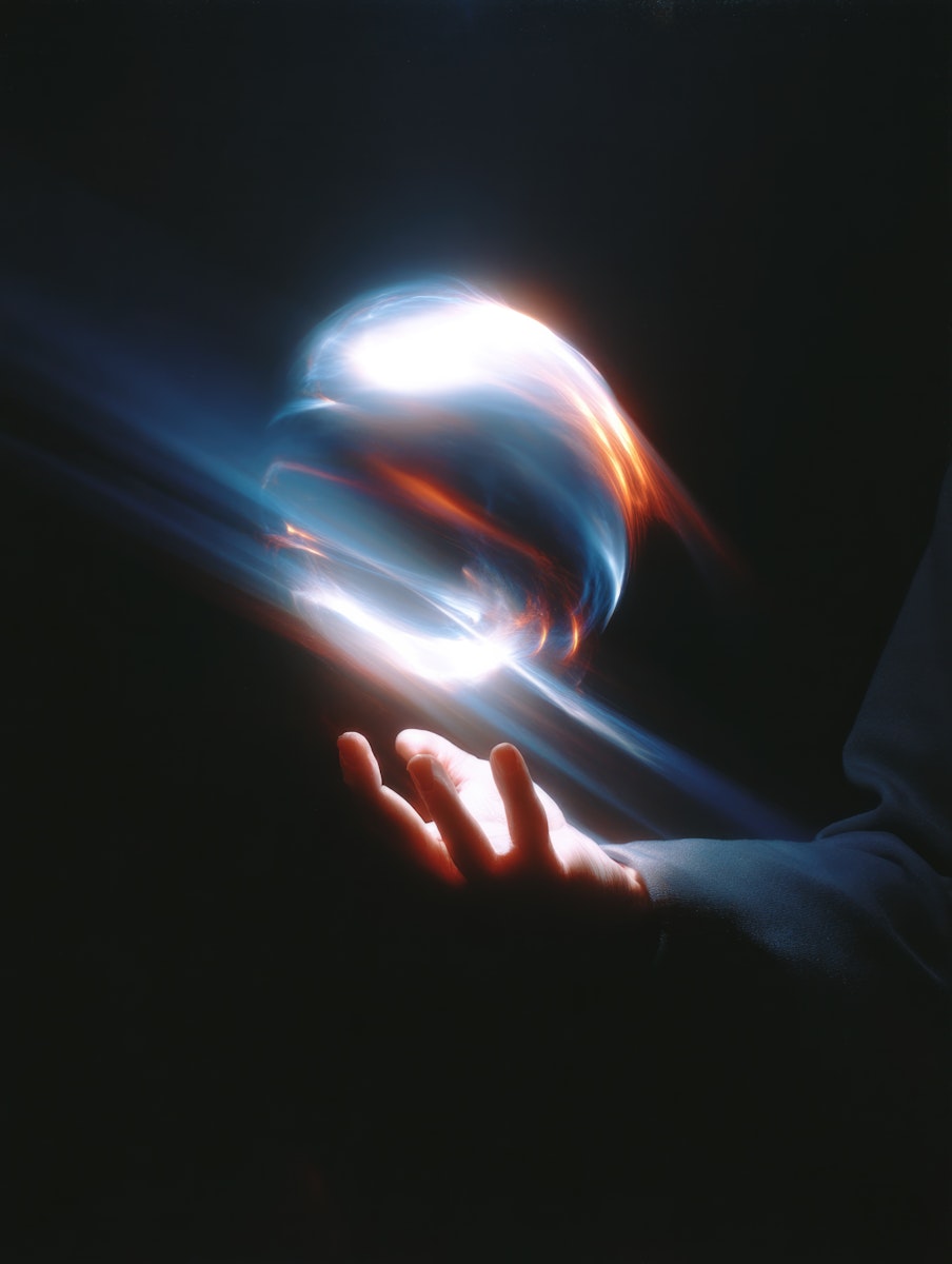 A glowing orb of light hovers above a hand in a dark setting, emitting vibrant streaks of blue, white, and orange light.