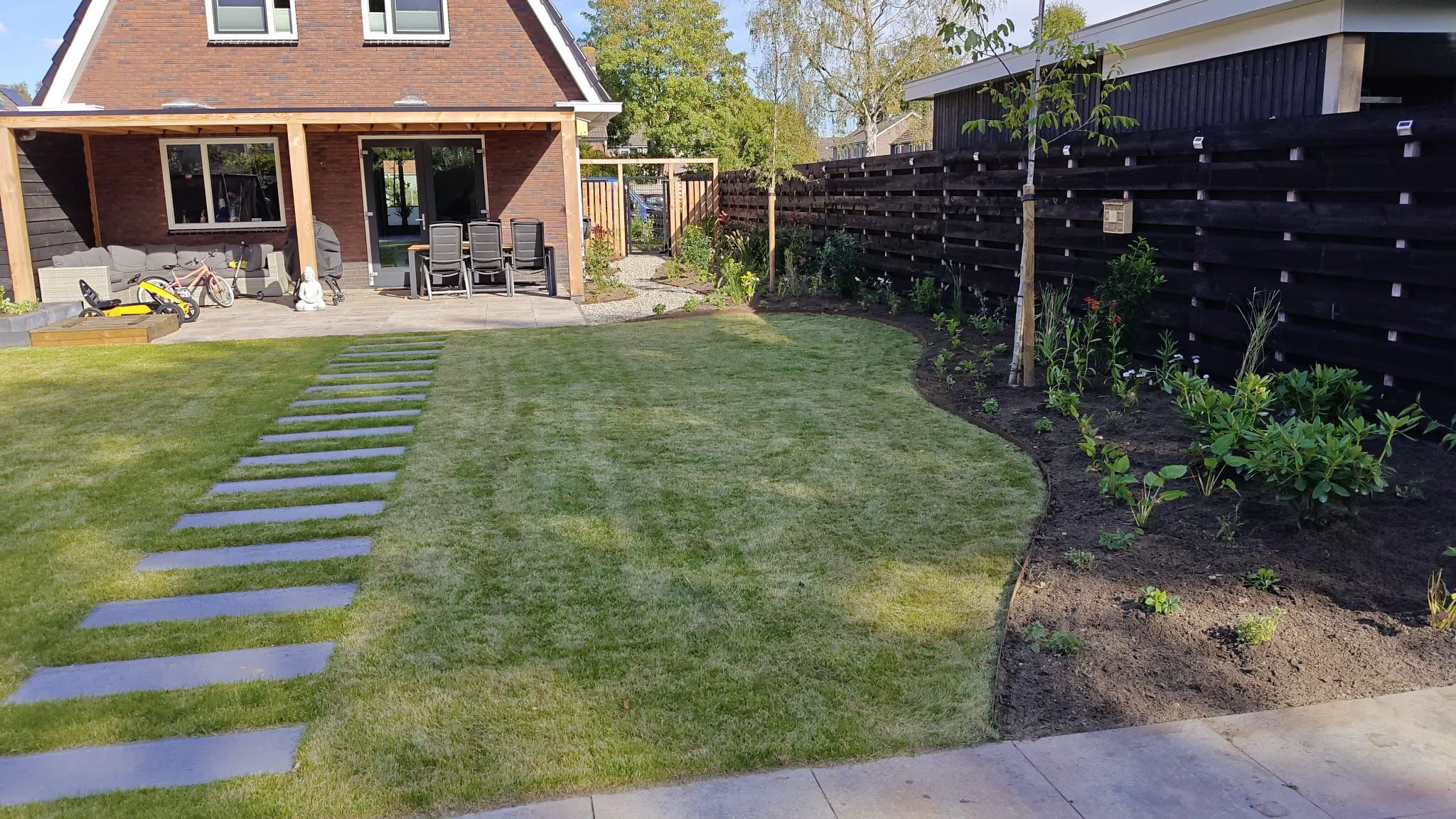 Strakke, kindvriendelijke tuin met nieuw gazon, bestrating, jonge beplanting en houten schutting rondom vrijstaande nieuwbouwwoning.