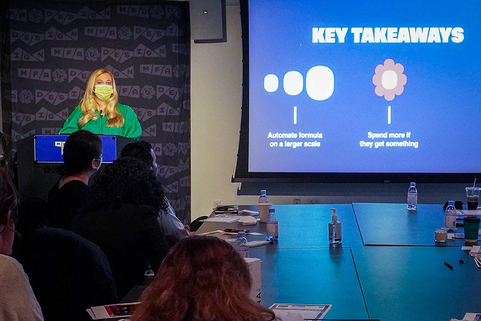 A woman dressed in green is giving a speech at the stand with an image projected on the right side of the wall with text and four graphical elements
