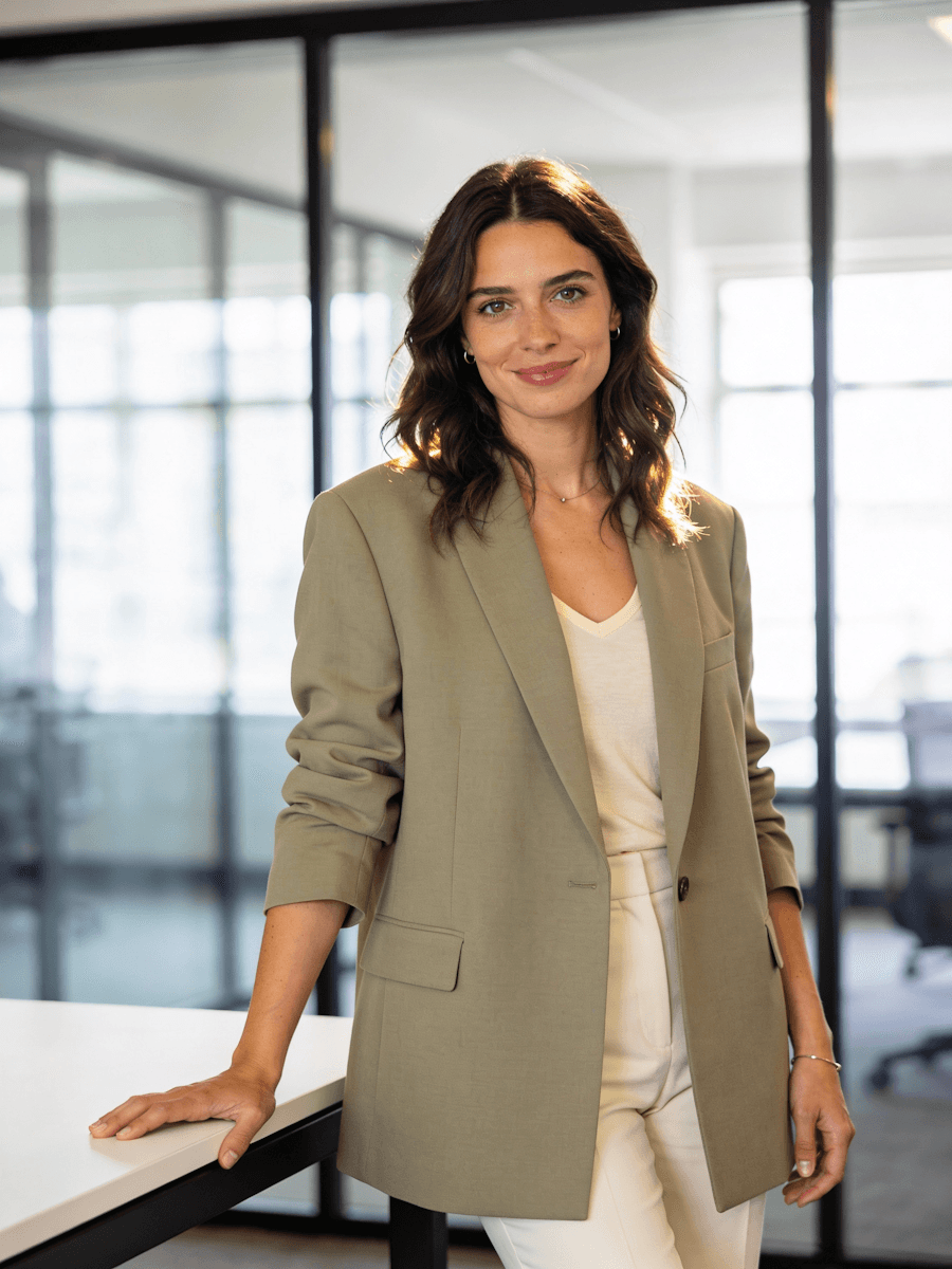 A woman in a light blazer stands confidently in a modern office setting with large windows.