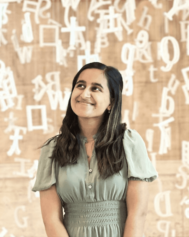 Geetanshi, an Indian American woman with long dark hair, stands in front of an art exhibit of floating letters. She is wearing a light green dress and is looking away from the camera.