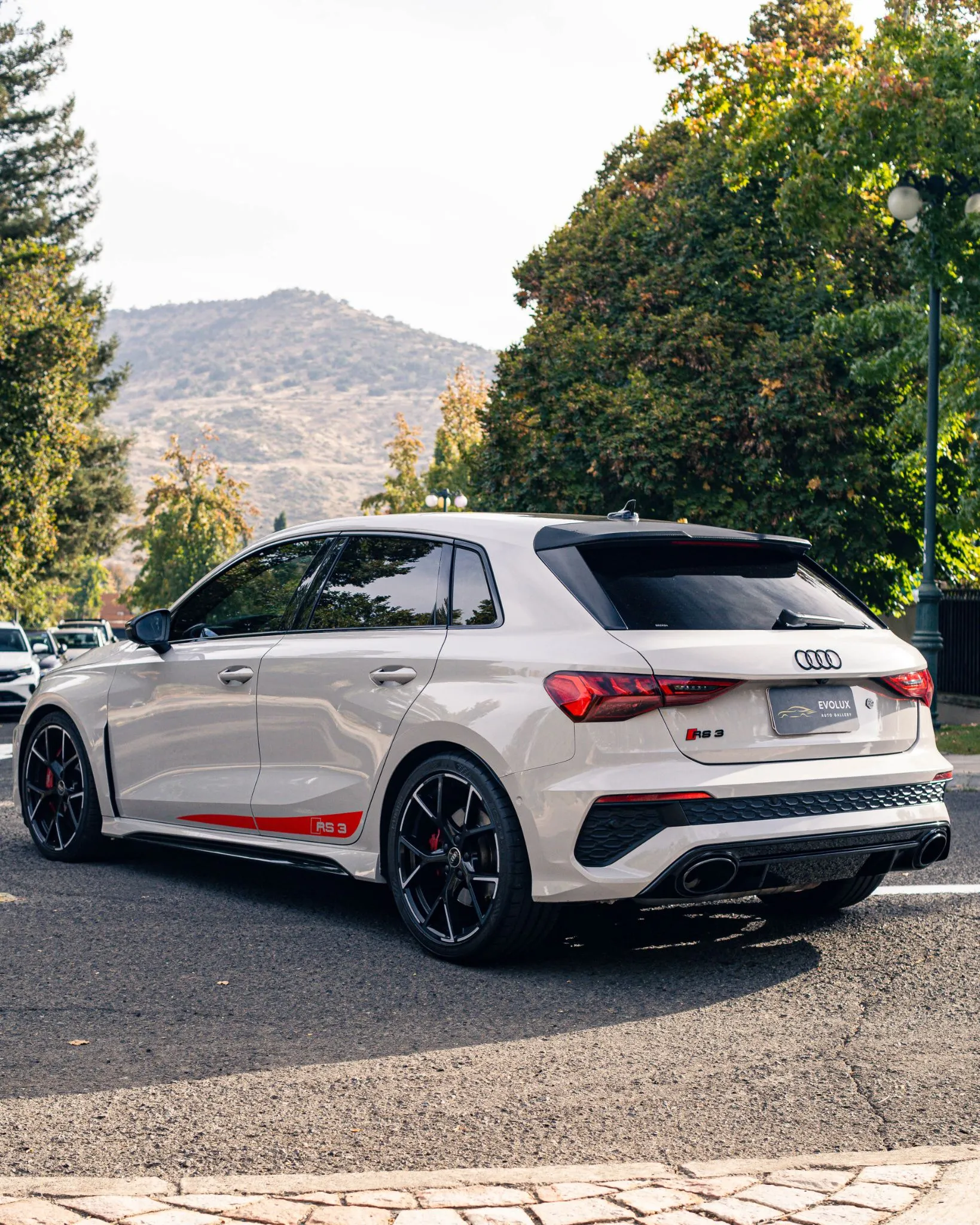 Audi RS3 Sportback blanco estacionado en calle arbolada, vista trasera lateral con llantas negras y emblema RS3