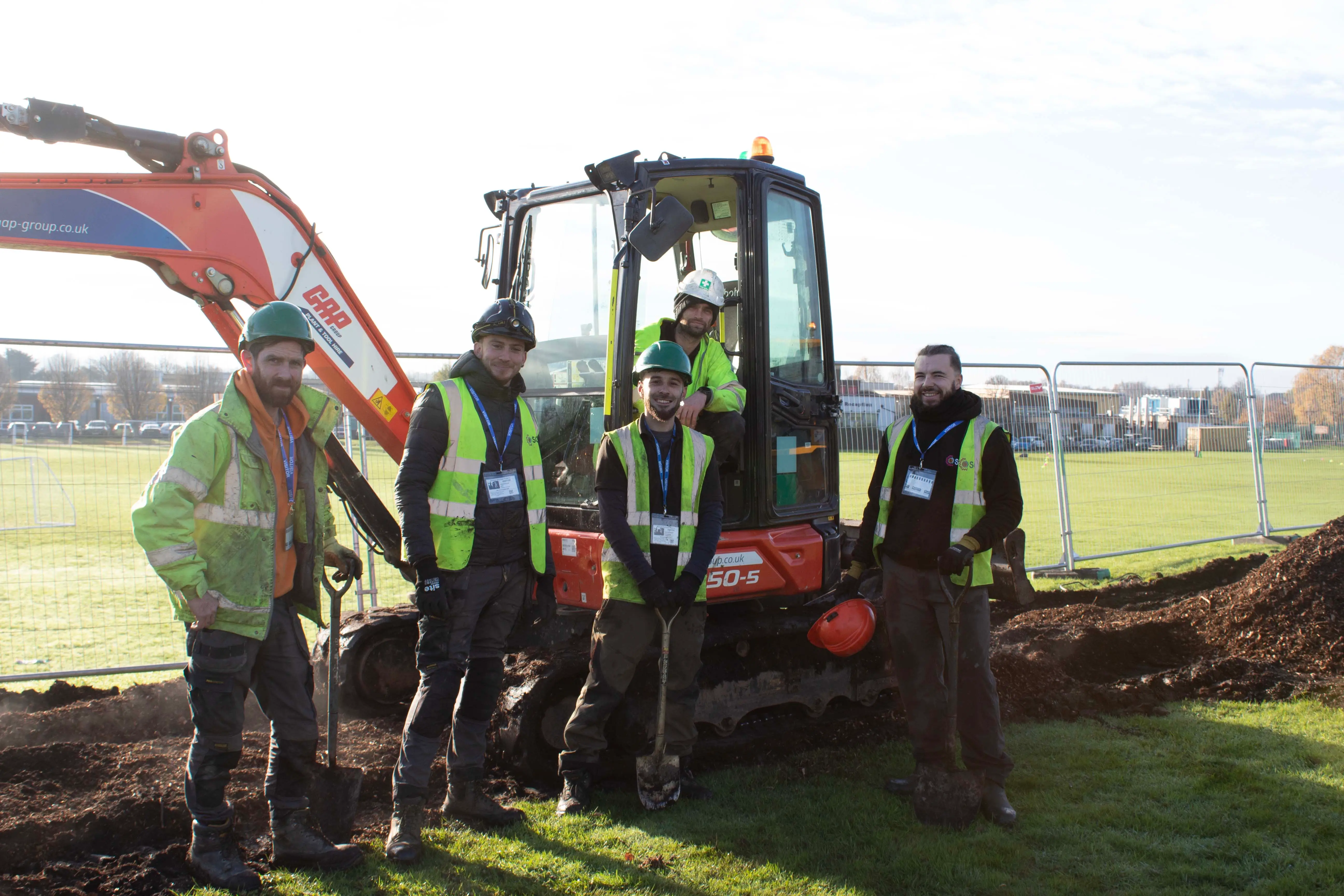 Construction workers operate machinery while digging in a grassy area under a clear sky.