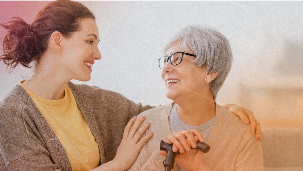 A compassionate caregiver in a mustard yellow shirt and brown cardigan smiles warmly at an elderly woman with silver hair and glasses, who is holding onto a walking cane and looking back with a joyful expression, indicative of trust and comfort.