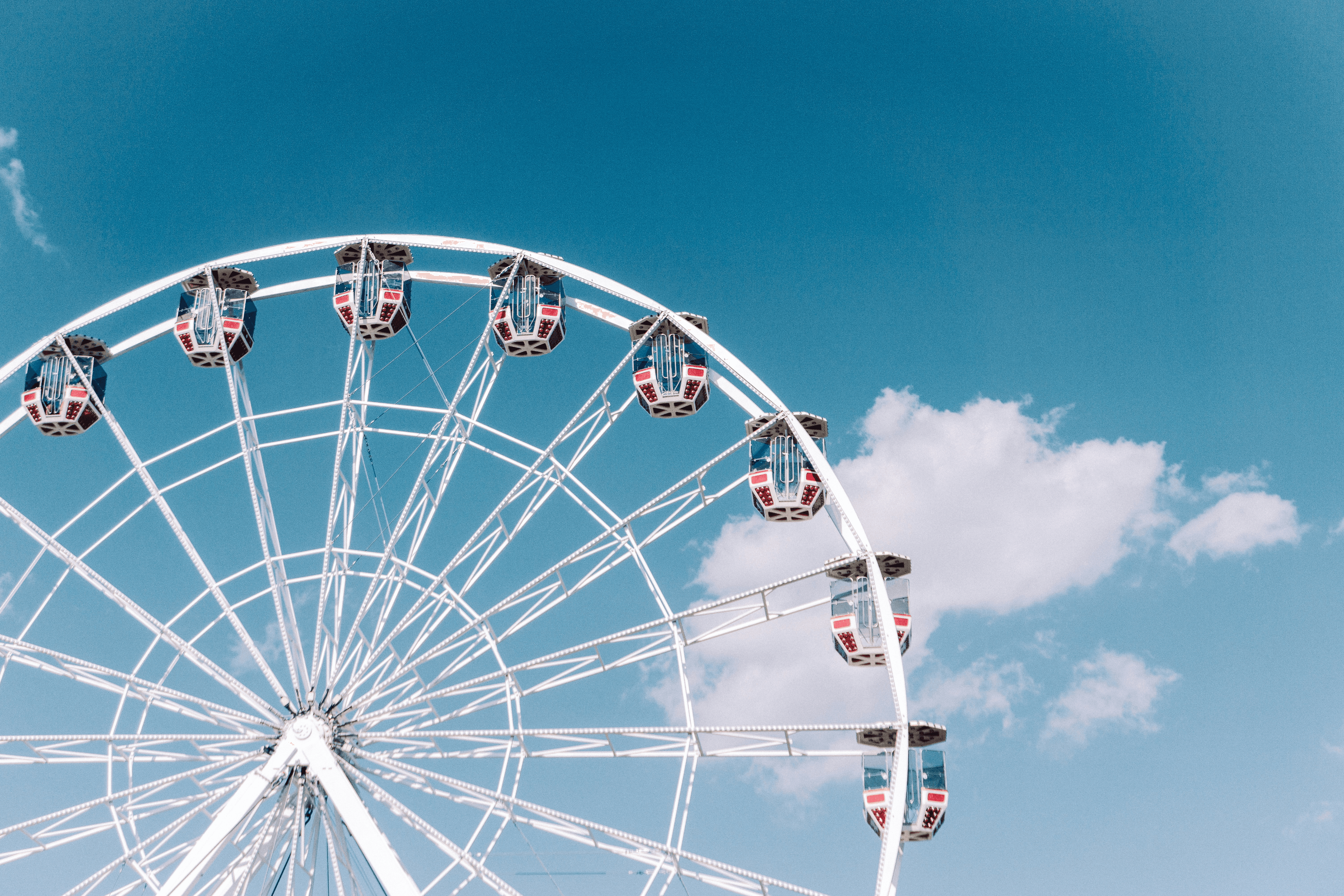 A low-angle shot of a white Ferris wheel with red and blue gondolas against a bright blue sky with a few white clouds.