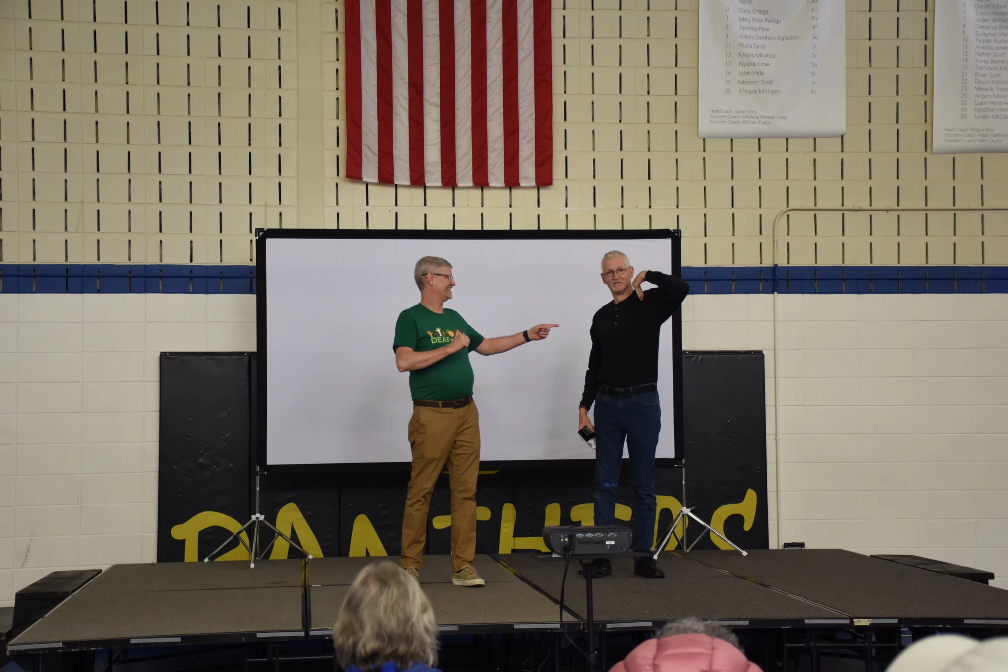 Two men giving a presentation on a stage in a gymnasium. One man is pointing. An American flag is visible on the wall.