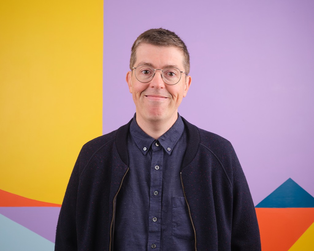 Portrait of Brian Sholis standing in front of a colourful mural