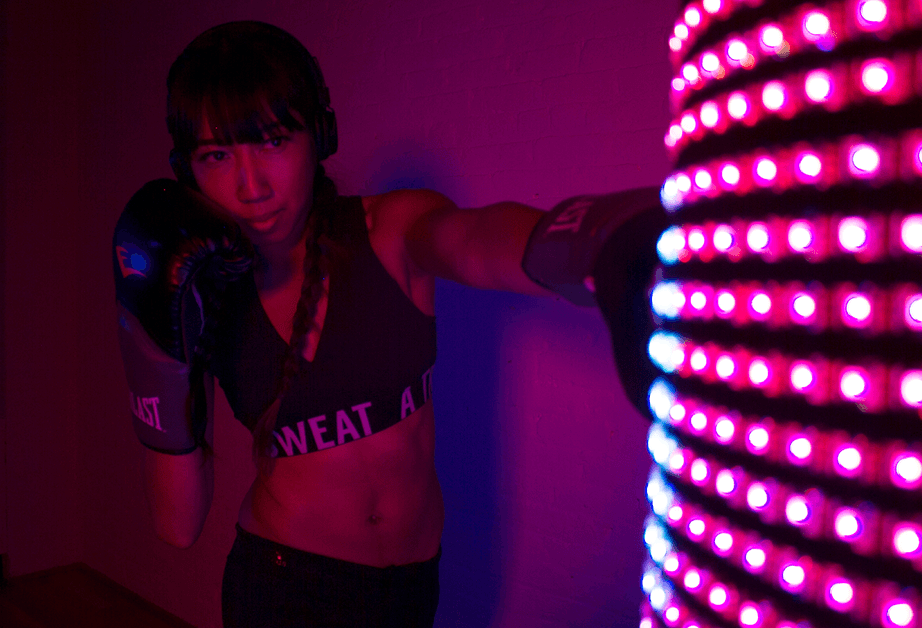 A young woman with dark hair pulled back in a braid wears a black sports bra with "WEAT A" printed on it and black shorts. She is wearing black boxing gloves and stands with her right arm extended toward a vertical display of red LED lights arranged in a grid pattern. The background is a solid purple wall.