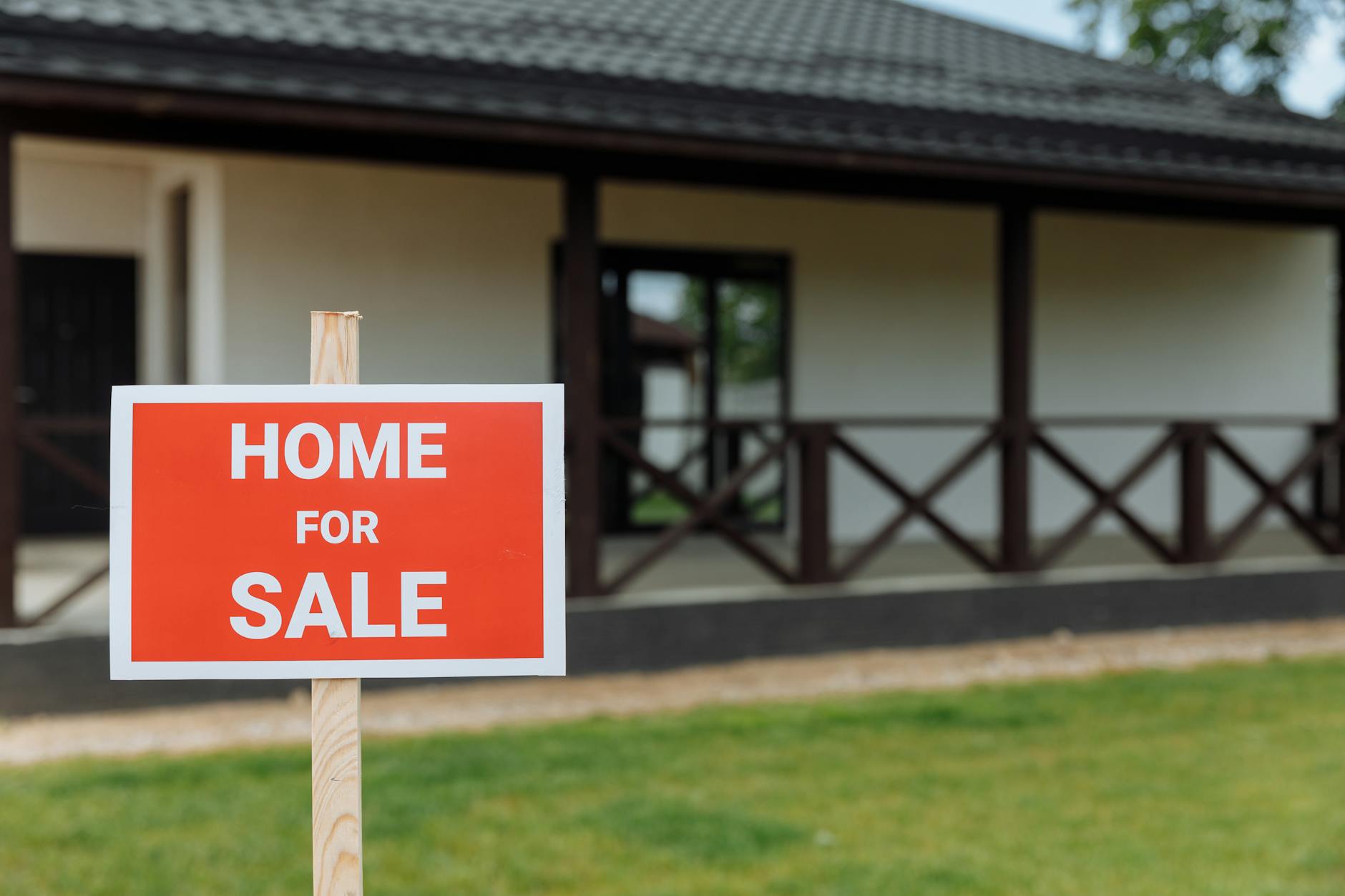 Red and white Home For Sale sign on a wooden post in front of a white bungalow with a covered veranda and green lawn