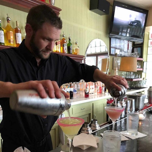 酒吧的調酒師從搖酒器中倒出兩杯雞尾酒，背景有展示的酒瓶和一台電視。