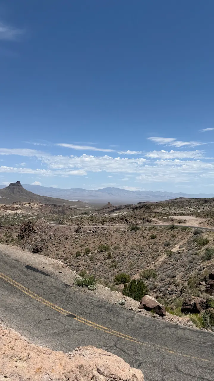 A winding desert road with blue sky, scattered clouds, and mountains in the distance.