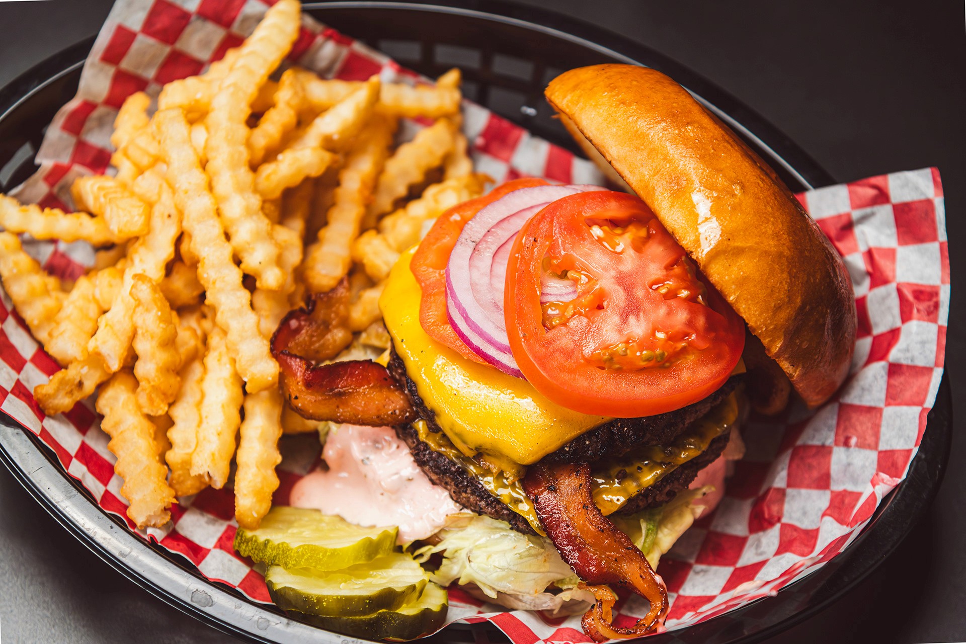 Gourmet cheeseburger with a grilled beef patty, melted cheese, sliced red onion, tomato, and lettuce on a brioche bun, served with a side of french fries in a plastic basket.