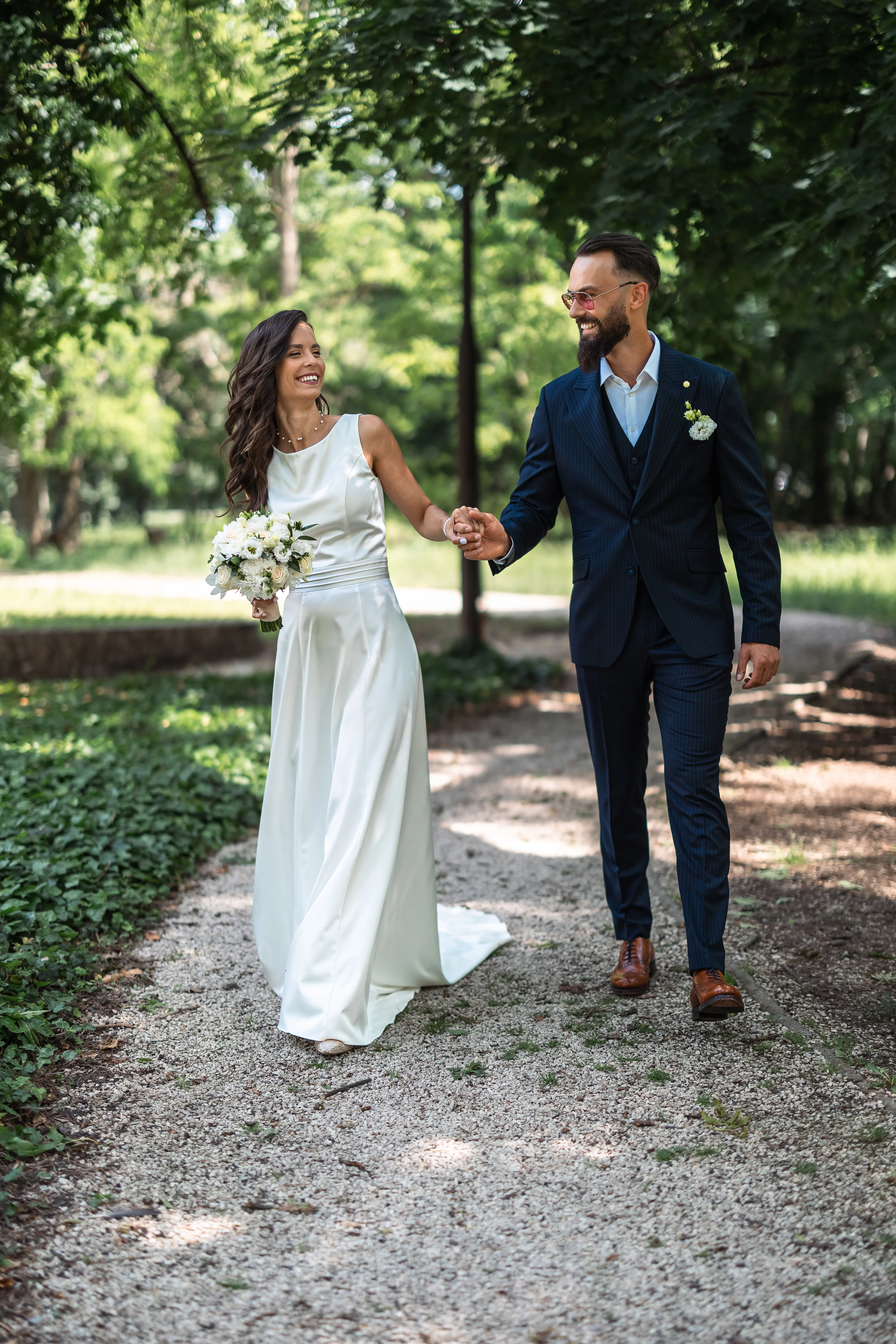 Boldog menyasszony fehér ruhában és vőlegény kék öltönyben kézen fogva sétálnak kavicsos parkúton a Fenyőharaszti Kastélyszálló fái között