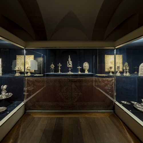 Glass display cases showcasing silver religious artifacts on blue velvet, illuminated with soft lighting in a museum setting.