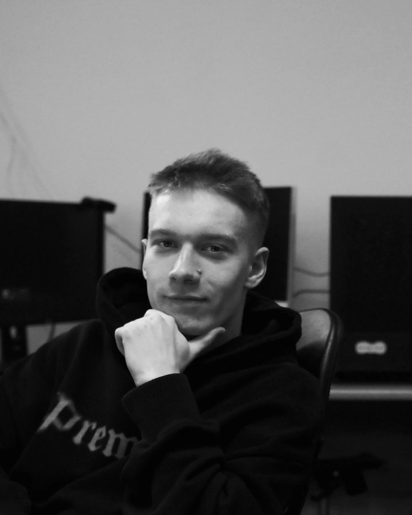 A black and white portrait of a young man with a slight smile, resting his chin on his hand while sitting in front of computer monitors.