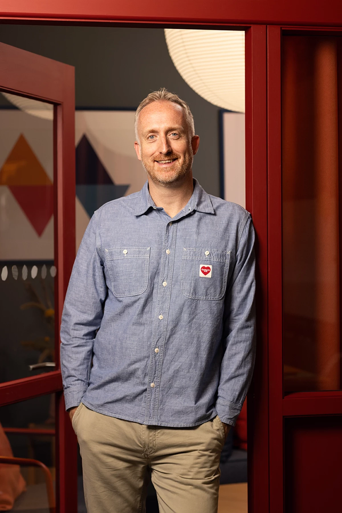 A man in a blue shirt and beige trousers stands in a red doorframe smiling at the camera